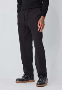Pantalon noir à coupe décontractée, taille élastique et poches latérales, accompagné de chaussures noires à lacets avec une semelle marron.
