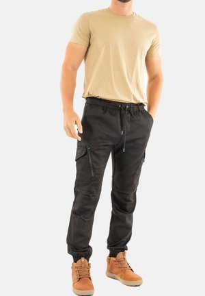 Pantalones cargo negros con puños elásticos y cintura con cordón; cuenta con bolsillos laterales con cremallera; se lleva con una camiseta beige y botas marrones.
