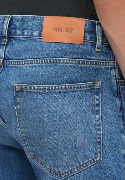 Kék farmernadrág bőrből készült "NN. 07" márkajelzéssel a hátsó derékrészen, klasszikus öt zsebes dizájnnal és kontrasztos varrással.