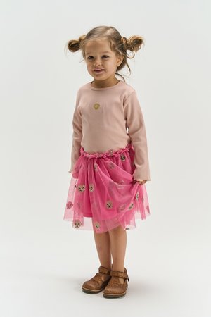 Jeune fille avec deux chignons portant un haut à manches longues rose clair, une jupe en tulle rose à motifs de coquillages, et des chaussures marron pailletées, debout.