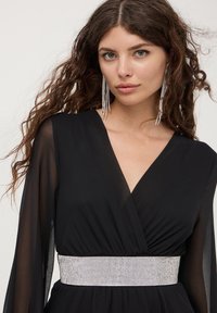 Abito nero avvolgente con maniche lunghe trasparenti e una cintura decorata con gioielli argentati. Capelli ricci e orecchini lunghi completano il look.