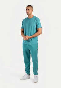 Grönblå bomulls-t-shirt med rund hals och korta ärmar, kombinerad med matchande grönblå byxor med fickor och avslappnad passform. Vita sneakers.