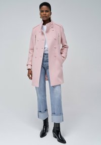 Abrigo doble faz de color rosa claro con hombreras y detalles de cremallera, llevado sobre una blusa blanca y jeans azul claro, combinado con botas negras de tacón.