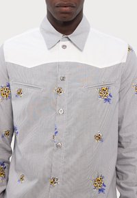 Camicia a righe con colletto e spalla bianchi; presenta ricami floreali gialli e blu; bottoni sulla parte anteriore; due tasche sul petto.