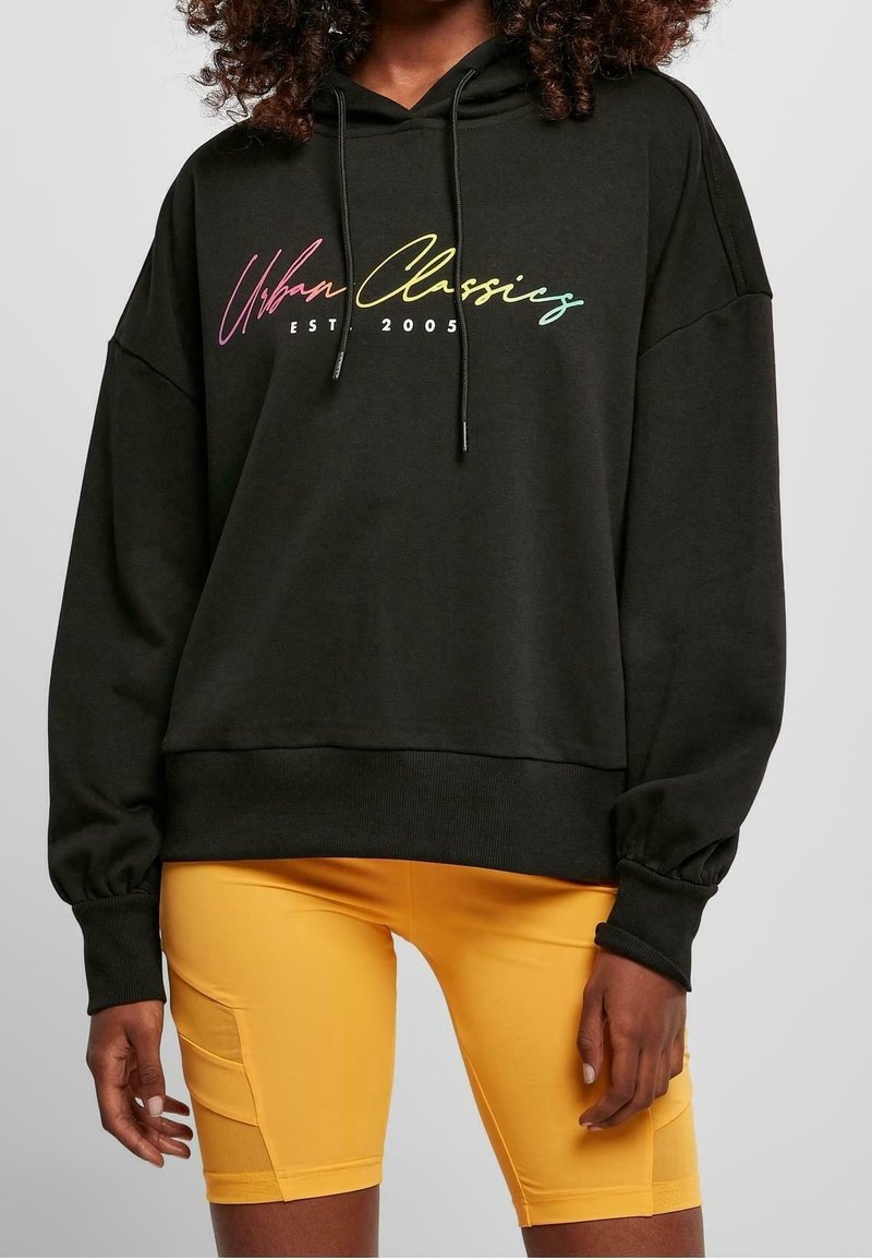 Černá oversized mikina s kapucí a šňůrkou, zdobená barevným logem "Urban Classics EST 2005". V kombinaci s žlutými přiléhavými šortkami.
