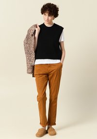 Czarny dzianinowy top bez rękawów na białej koszulce, brązowe spodnie, beżowe buty wsuwane, trzymając kurtkę w panterkę.