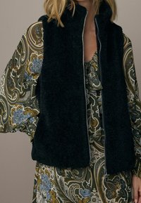 Zwart, pluizig vest met hoge kraag en volledige ritssluiting, gedragen over een kleurrijke jurk met een paisley-patroon in groen, blauw en bruine tinten.