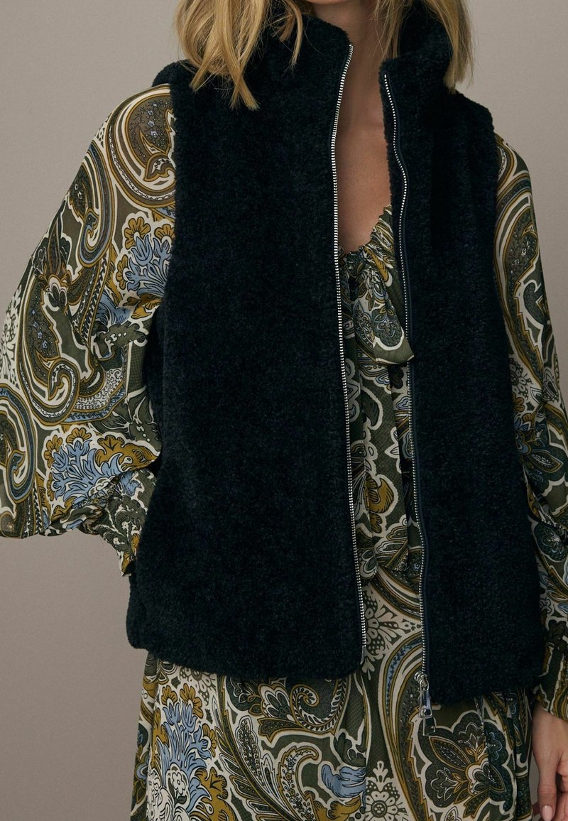 Zwart, pluizig vest met hoge kraag en volledige ritssluiting, gedragen over een kleurrijke jurk met een paisley-patroon in groen, blauw en bruine tinten.