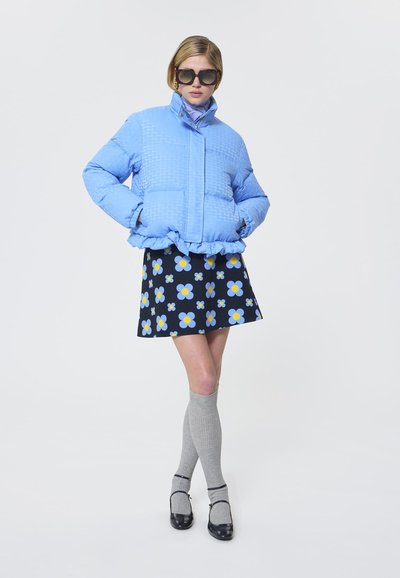 Jachetă puffer căptușită de culoare albastru deschis, cu tiv ondulat, asortată cu o fustă neagră cu modele de flori galbene și albastre, sosete gri până la genunchi și pantofi negri.