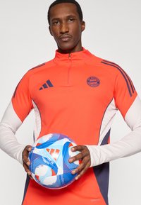 Rød atletisk zip-up genser med marineblå detaljer, med FC Bayern München-logo. Holder en blå og hvit mønstret fotball.