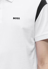Polo blanc avec une bande noire à l'épaule et le logo noir "BOSS" brodé sur la poitrine, porté par une personne avec un bras nu visible.