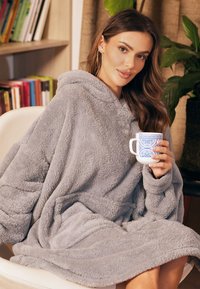 Szary polarowy koc-hoodie w oversize'owym kroju o miękkiej fakturze, z kapturem i kieszeniami. Osoba trzyma niebieską ceramiki kubek w wzory.