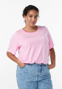 Ung kvinde med krøllet hår iført en lyserød og hvid stribet T-shirt og lyseblå bukser med hjerte mønstre, hænderne i lommerne, smilende.