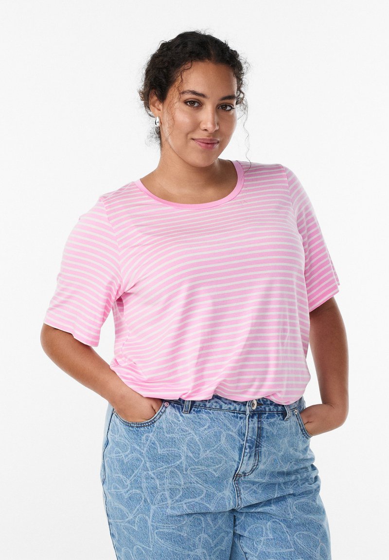 Ung kvinde med krøllet hår iført en lyserød og hvid stribet T-shirt og lyseblå bukser med hjerte mønstre, hænderne i lommerne, smilende.