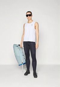 Balti sportinė be rankovių palaidinė, juodos tamprės su šoninėmis kišenėmis ir tamsūs batai. Rankose šviesiai mėlynas kuprinės su mėlynomis detalėmis.