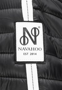 Sort polstret stof med et hvidt label med "NAVAHOO"-logoet, etableret i 2014, og en kontrasterende stribet accent.