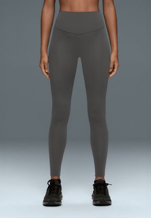 Femme portant un legging gris foncé taille haute et des chaussures de sport noires, debout devant un fond gris uni.