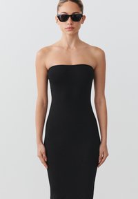 Juodas strapless bodycon suknelė, pagaminta iš prigludusios medžiagos, lygaus paviršiaus, iki kelių ilgio. Aksesuaru naudojamos saulės akiniai su juodais rėmeliais.
