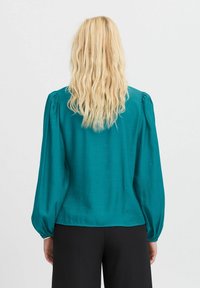 Blusa teal con maniche lunghe a sbuffo, texture liscia e vestibilità morbida. Il orlo si ferma in vita, con polsini elastici.