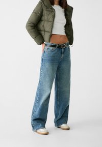 Olivgrön vadderad jacka, vit crop top, vida blå jeans med svart bälte och beiga sneakers med vita detaljer.