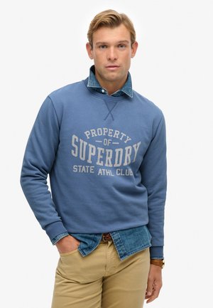 Felpa blu con stampa "PROPERTY OF SUPERDRY", indossata sopra una camicia di jeans. Presenta polsini a coste e una vestibilità comoda. Dettagli color crema.