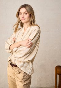 Femme aux cheveux châtain clair portant un chemisier beige rayé et un pantalon beige clair, debout les bras croisés devant un mur clair uni.