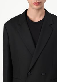 Black blazer with a structured silhouette, notch lapels, and double-button closure, paired with a black crew neck shirt underneath.