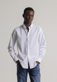 Joven de piel oscura vestido con una camisa blanca de botones y jeans azules, de pie con las manos en los bolsillos frente a un fondo sencillo.