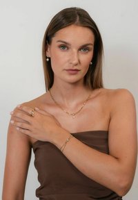 Gold jewelry set featuring a chain necklace with a pearl pendant, stud earrings, a delicate bracelet, and a ring. Worn on a model with bare shoulders.