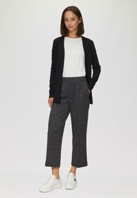 Zwarte cardigan over een witte T-shirt, gecombineerd met grijze broek met patroon en witte sneakers. Losse pasvorm, met zakken in de broek.