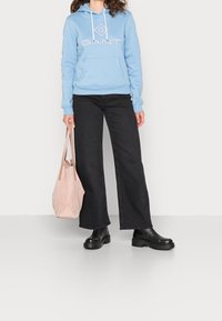 Lichtblauwe hoodie met een logo, voorkantzak en trekkoord. Gecombineerd met zwarte wijdvallende jeans en chunky zwarte enkellaarsjes. Houdt een roze tas vast.