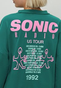 Zeleni dukserica s upadljivim ružičastim natpisom "SONIC RADIO US TOUR." Uključuje razigrane ružičaste siluete i popis datuma turneje u bijeloj boji.