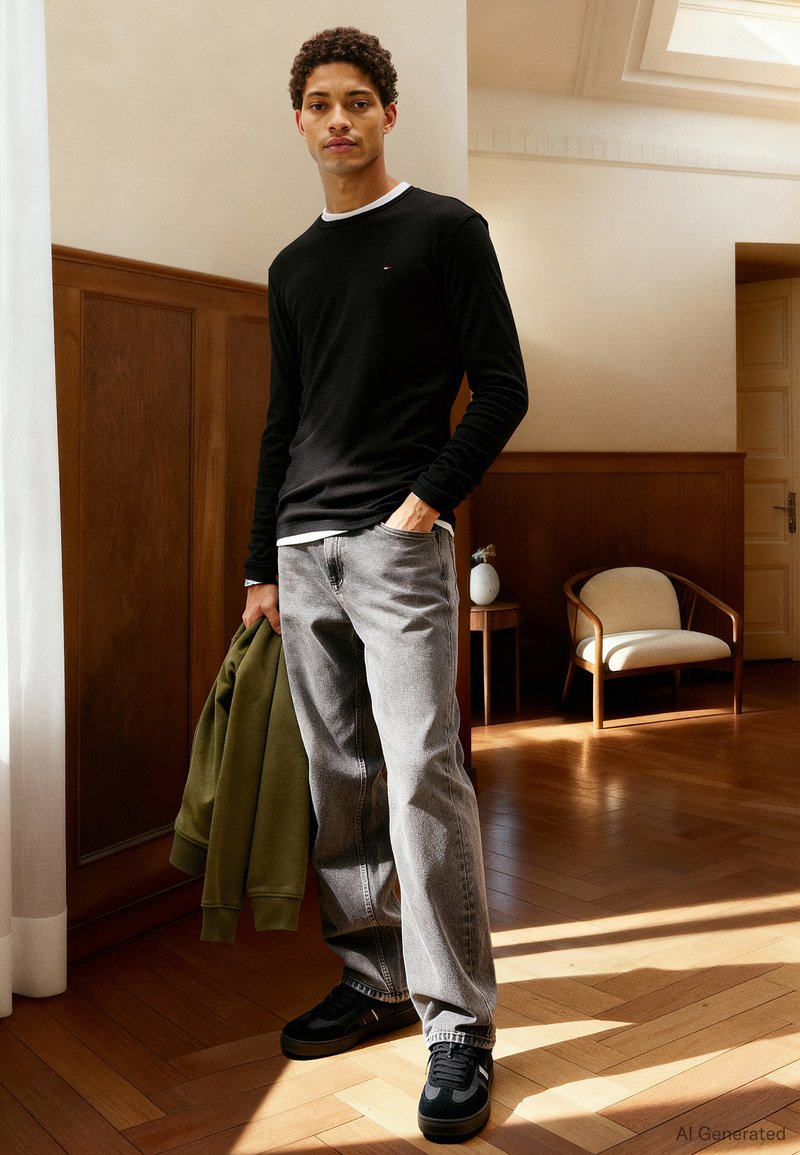 Junger Mann in schwarzem Langarmshirt, grauer Jeans, der eine grüne Jacke hält, steht in einem sonnenbeschienenen Raum mit Holzvertäfelung und einem Sessel.