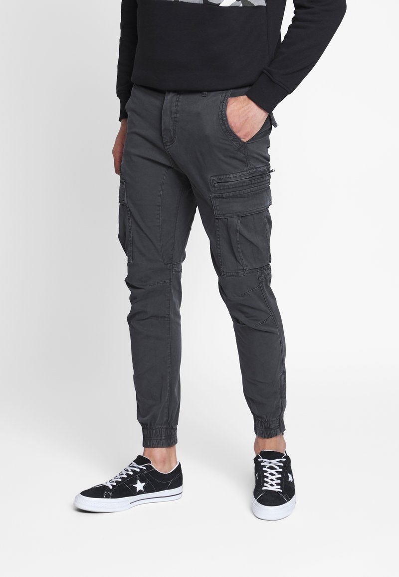 Homme portant un pantalon cargo gris foncé avec des poignets élastiques et des baskets basses noires avec un détail étoile blanche, debout avec une main dans la poche.