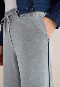 Gros plan d'une personne portant un pantalon de survêtement gris avec cordon de serrage et une main dans une poche latérale, associé à une veste bleu marine avec des poignets élastiques.