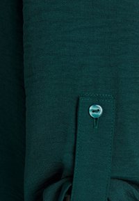 Tessuto testurizzato color turchese con dettaglio a bottone. Il tessuto presenta un motivo intrecciato sottile, evidenziando una finitura morbida e leggera e una texture liscia.