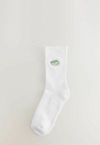 Vitt bomullssocka med ribbad textur, försett med en cirkulär broderad lapp i gröna och rosa nyanser, placerad vid vadens mitt.