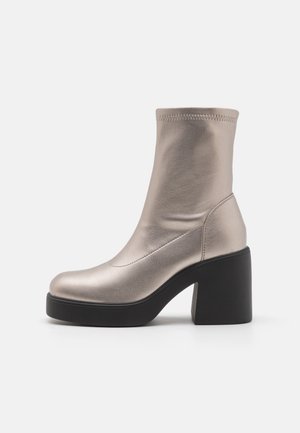 Bottine argentée métallisée avec une texture en cuir lisse, bout arrondi et un talon épais noir. Présente un tige ajustée et élastique.
