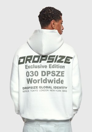 Mężczyzna w białej bluzie z kapturem na głowie, z tyłu wyraźny ciemny napis: „DROPSIZE Exclusive Edition 030 DPSZE Worldwide” oraz nazwy miast.