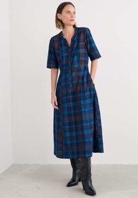 Egy nő kék és barna kockás midi ruhát visel félhosszú ujjakkal és fekete magassarkú csizmával, egy sima fehér fal előtt áll.