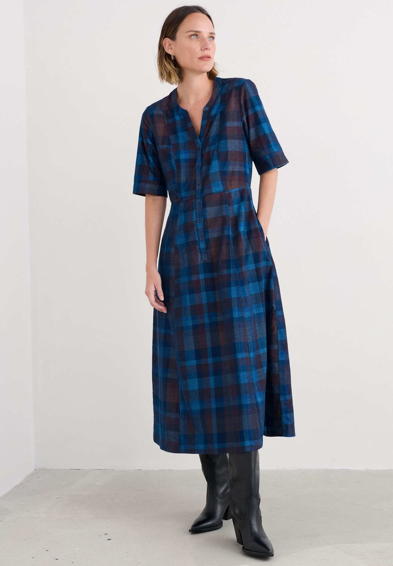 Egy nő kék és barna kockás midi ruhát visel félhosszú ujjakkal és fekete magassarkú csizmával, egy sima fehér fal előtt áll.