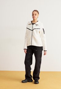 Jaqueta de fleece branca com detalhes pretos, fecho de zip e bolso lateral, combinada com calças largas pretas e ténis pretos.
