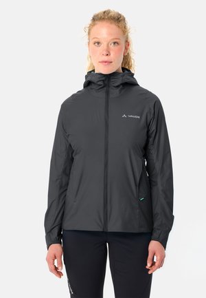 Femme de face portant une veste noire à capuche zippée et un pantalon noir sur un fond clair uni.