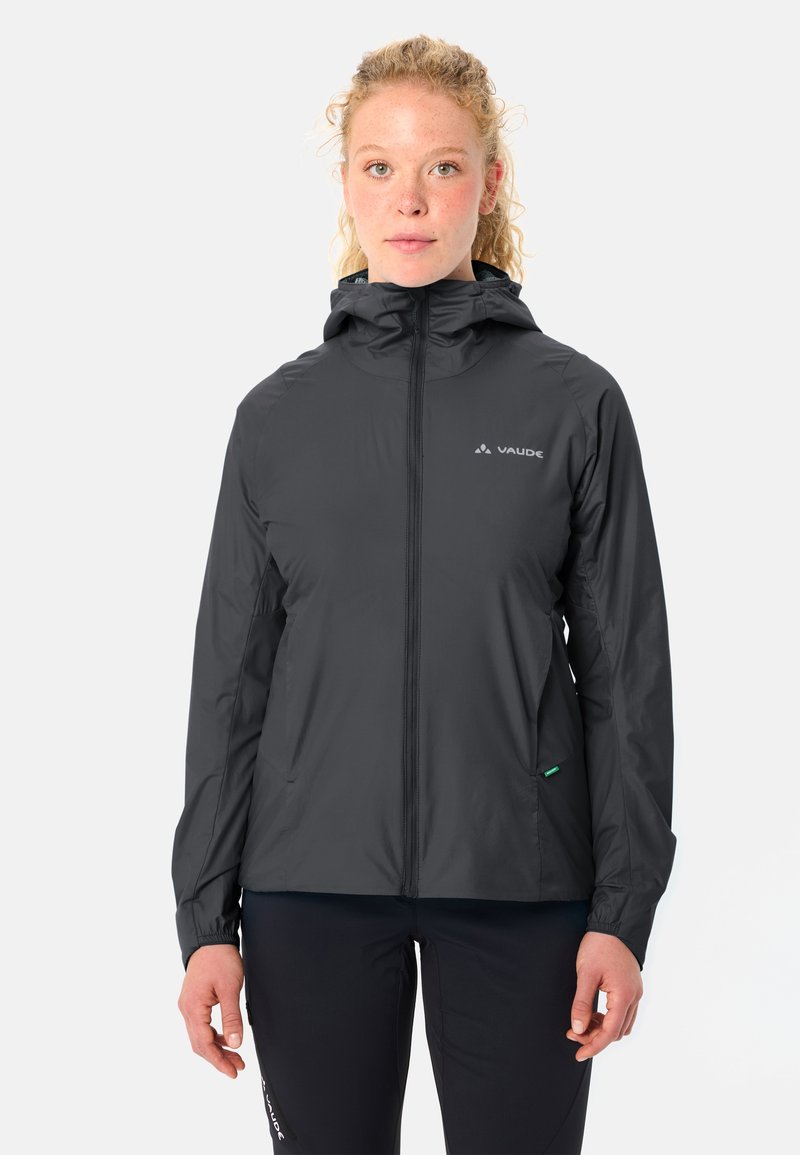 Femme de face portant une veste noire à capuche zippée et un pantalon noir sur un fond clair uni.