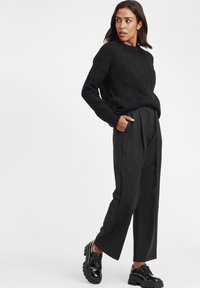 Maglione nero a trecce con maniche lunghe, abbinato a pantaloni neri a vita alta con pieghe e scarpe basse nere con lacci spessi.