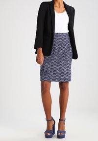 Frau in dunkelblauer, strukturiertem Bleistiftrock, weißem Oberteil, schwarzem Blazer und blauen High Heels, vor einem schlichten Hintergrund stehend.