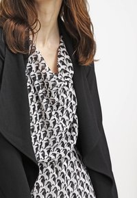 Svart jacka över en vit blus med ett mönster av svarta katter, med en draperad halsringning och en mjuk, flödande textur.