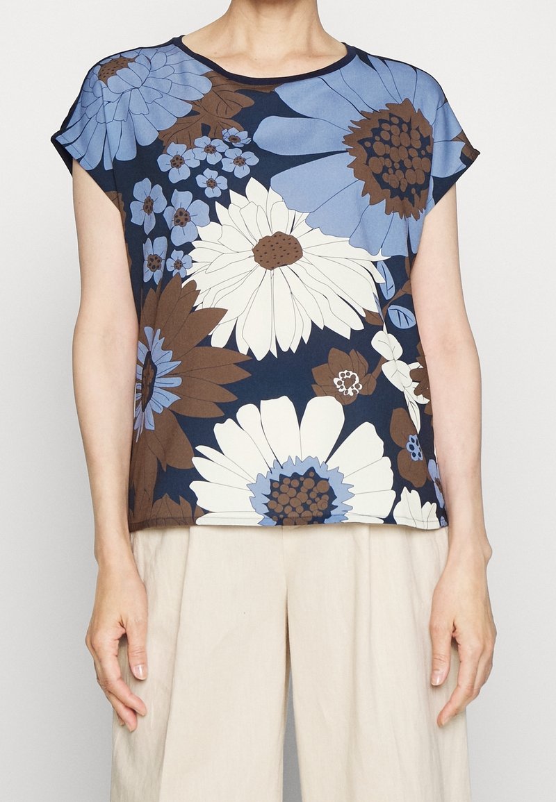 Haut floral bleu marine avec grandes fleurs blanches, bleues et marron porté avec un pantalon beige, visible du cou aux hanches.