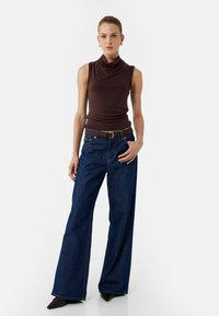 Ciemnobrązowy bezrękawnik z dekoltem typu cowl, zestawiony z szerokimi, niebieskimi dżinsami i brązowym paskiem; modelka nosi czarne szpilki.