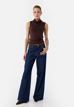 Haut sans manches marron foncé avec un col drapé, associé à un jean en denim bleu à jambe large et une ceinture marron ; le modèle porte des chaussures noires à talons pointus.